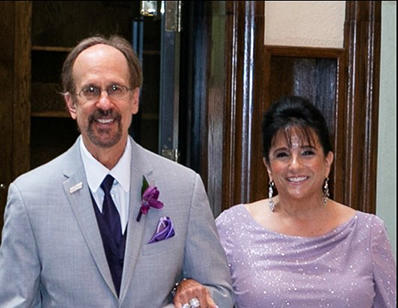Greg Baroni, with glasses and a goatee, is wearing a grey suit with a purple tie, boutonierre and pocket square. Camille Baroni wears a lavender gown.